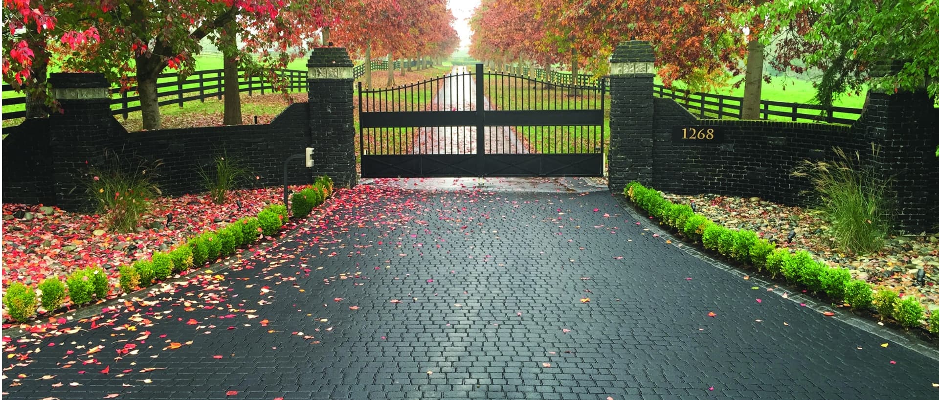 StreetPrint stamped asphalt estate driveway — dark cobblestone-pattern entry between brick gate pillars under autumn trees
