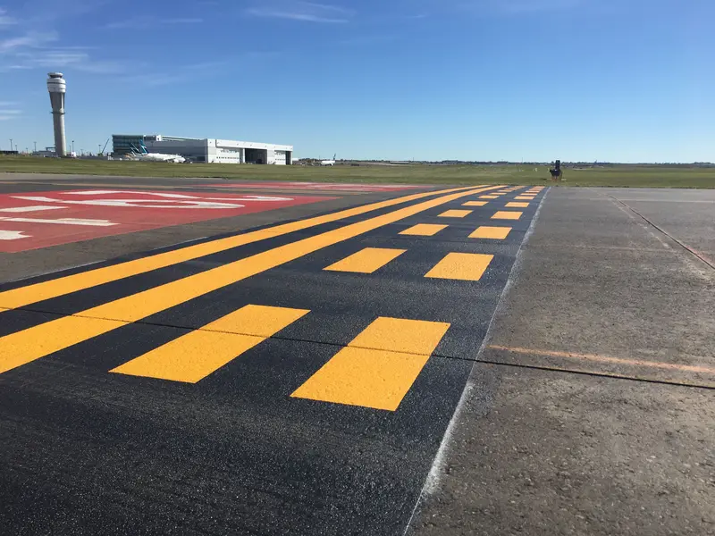Yellow AirMark holding-position markings on an airport taxiway, control tower behind